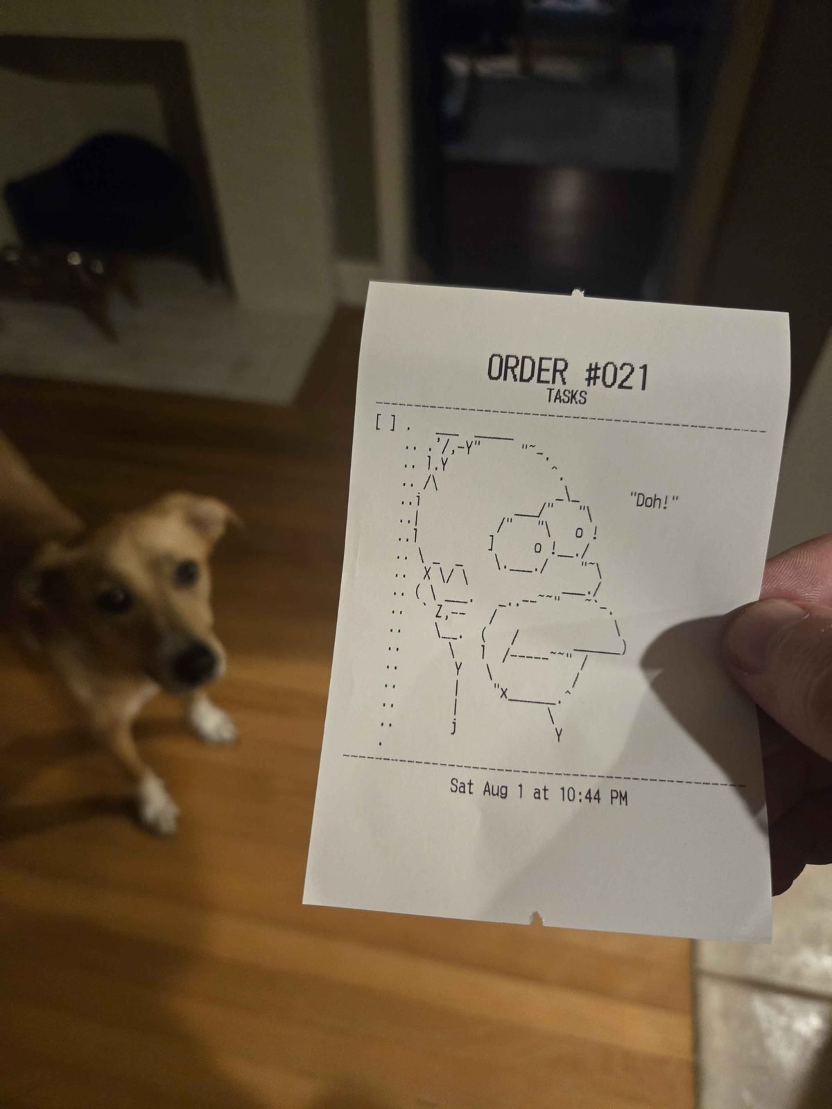 A printed receipt held up in front of a curious dog. The ticket reads ORDER #021 / TASKS and contains a large ASCII-art drawing made of punctuation characters.