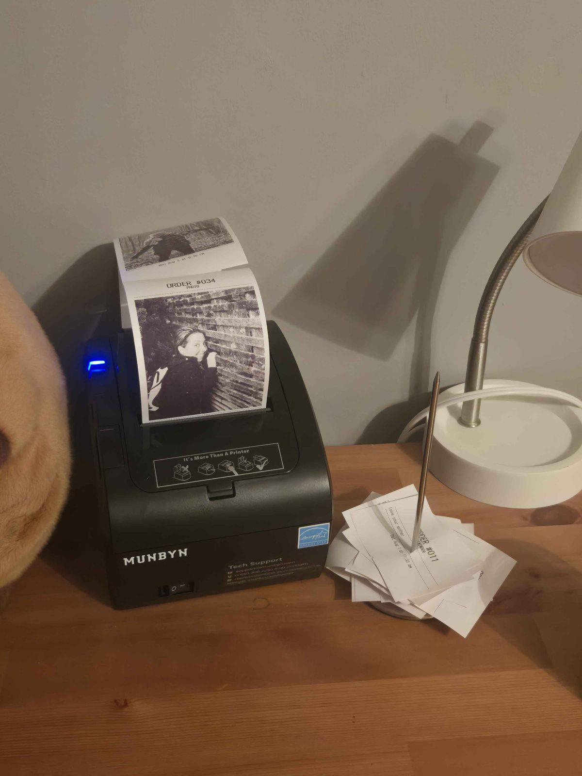 A MUNBYN thermal receipt printer on a desk, mid-print, with a ticket headed ORDER #034 / PHOTO emerging from it carrying a dithered black-and-white photograph. To the right, a metal spike holds a thick stack of finished tickets.